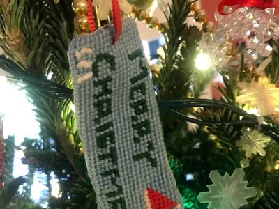 Blue cross-stitched ornament reading "MERRY CHRISTMAS" with a small red triangle on a tree