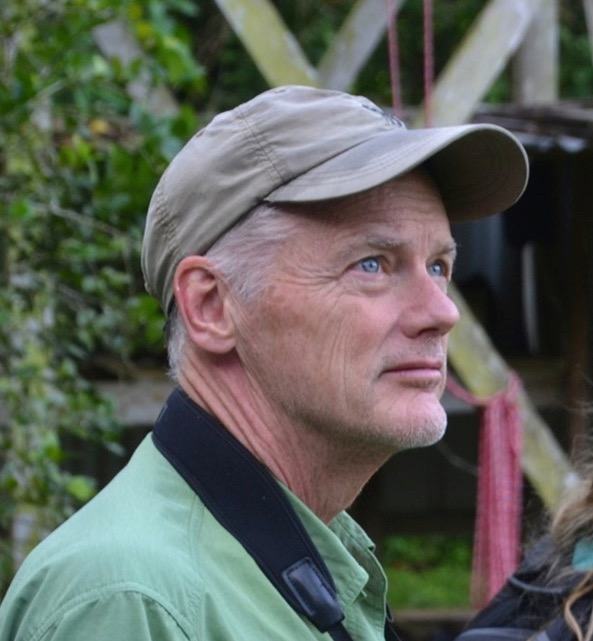 Man wearing a beige cap and green shirt, looking upward outdoors with camera strap visible
