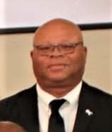 Bald man wearing glasses, dark suit, white shirt, black tie, small lapel pin