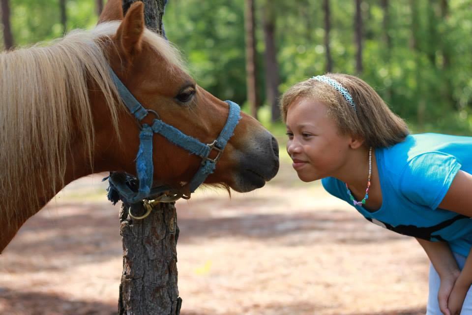 Girl leaning forward to touch noses with a small brown pony tied to a tree