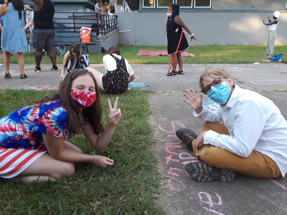 Two masked children on grass and sidewalk waving and showing a peace sign during chalk activity