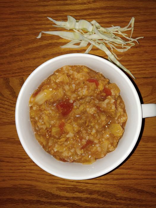 White mug of tomato‑rice and cabbage stew on wooden table, shredded cabbage beside it