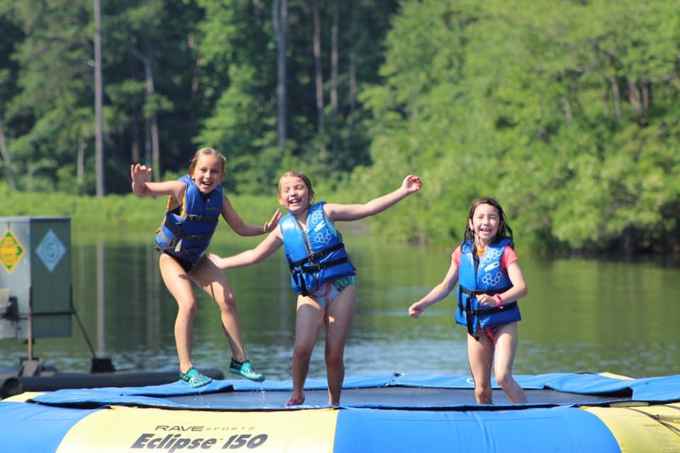 Three children in life jackets jumping on a water trampoline labeled Eclipse 150 on a lake