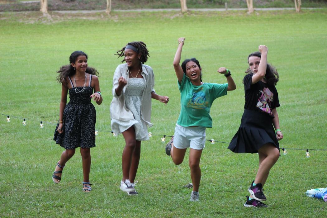 Four girls running and jumping on grass near a low string of lights