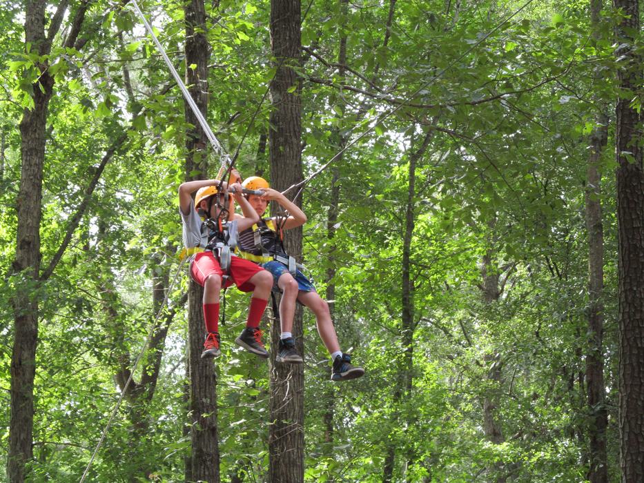 Two children in helmets suspended by harnesses on a ropes course among trees