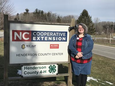 Person beside sign reading "NC Cooperative Extension, Henderson County Center; Henderson County 4‑H"