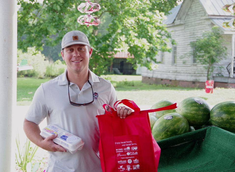 Man holding egg carton and red bag labeled "Enjoy local foods, visit local farms" beside watermelons