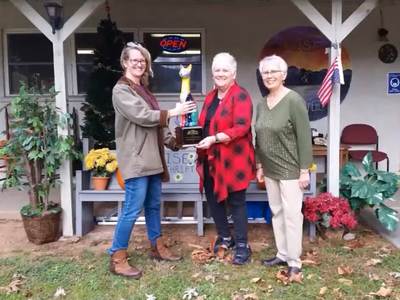 Three women holding a colorful cat-shaped trophy outside a thrift shop with "OPEN" sign.