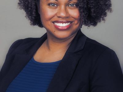 Smiling woman with curly hair wearing a blue top and black blazer, head-and-shoulders portrait