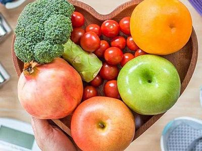 Hand holding heart-shaped bowl of fruits and broccoli; dumbbells, scale, yoga mat, shoes around
