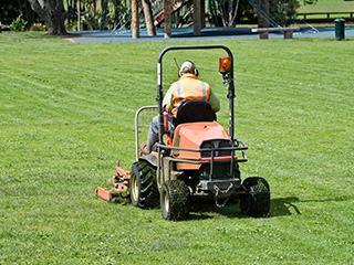 Worker in high-visibility vest on ride-on mower cutting grass on a lawn.