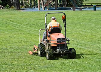 Worker in high-visibility vest on ride-on mower cutting grass on a lawn.