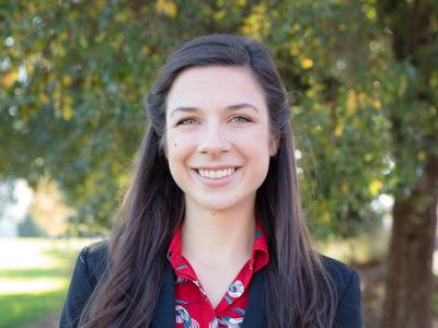 Woman smiling outdoors wearing red floral blouse and black blazer, tree background
