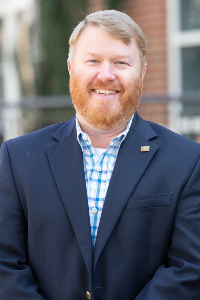 Smiling man with red beard and short blond hair wearing a navy blazer and blue plaid shirt