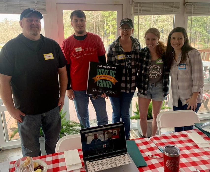 Five people standing indoors holding a "Participating Visit NC Farms Randolph County" sign