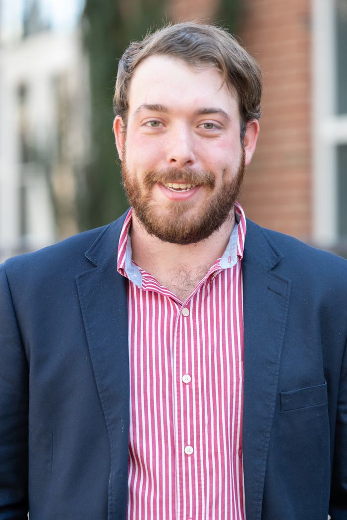 Young bearded man facing camera, wearing red-striped shirt and dark blazer