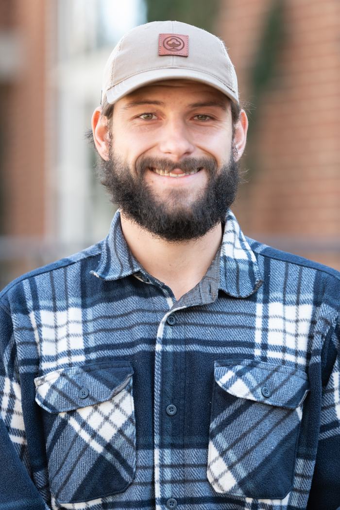 Bearded man wearing beige cap and blue plaid shirt, facing camera outdoors