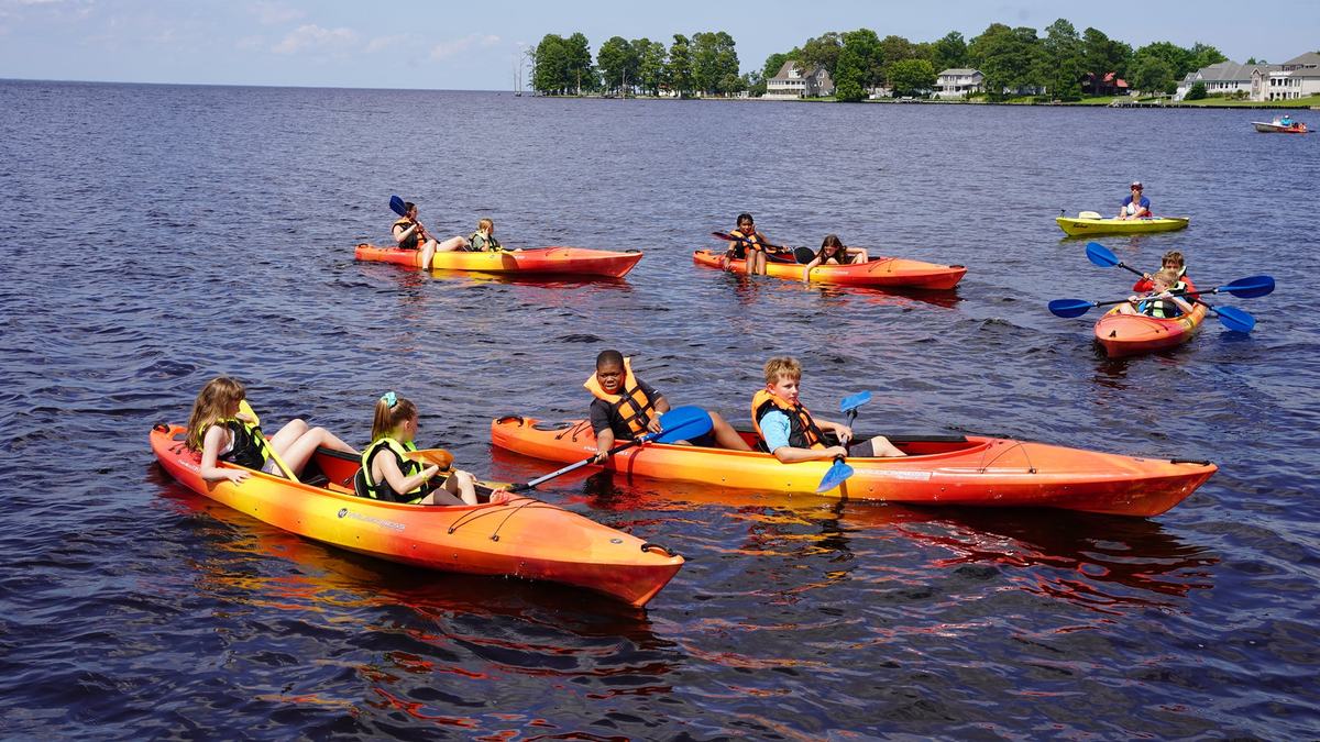 Children wearing life jackets kayaking in colorful kayaks on a lake near shoreline houses