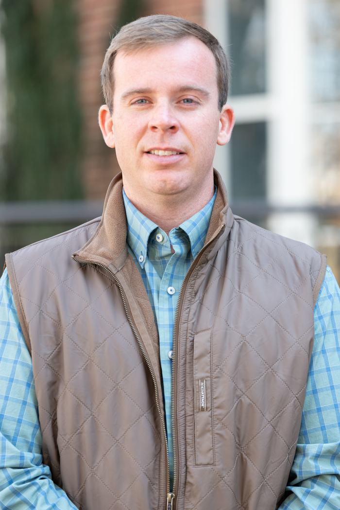 Man wearing brown quilted vest over blue checkered shirt, facing camera