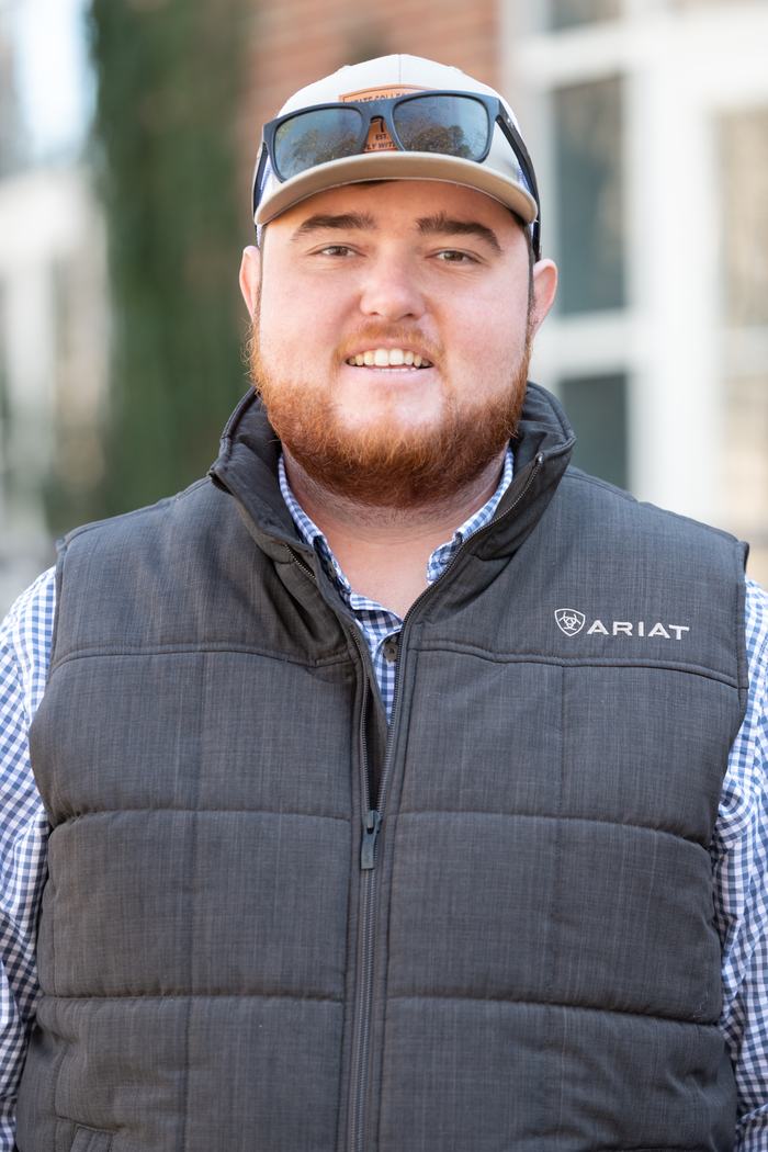 Young man wearing a cap with sunglasses on it, gray ARIAT vest, looking at camera