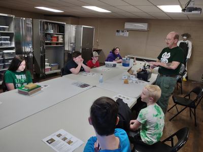 Instructor standing at table teaching children seated around tables with worksheets.