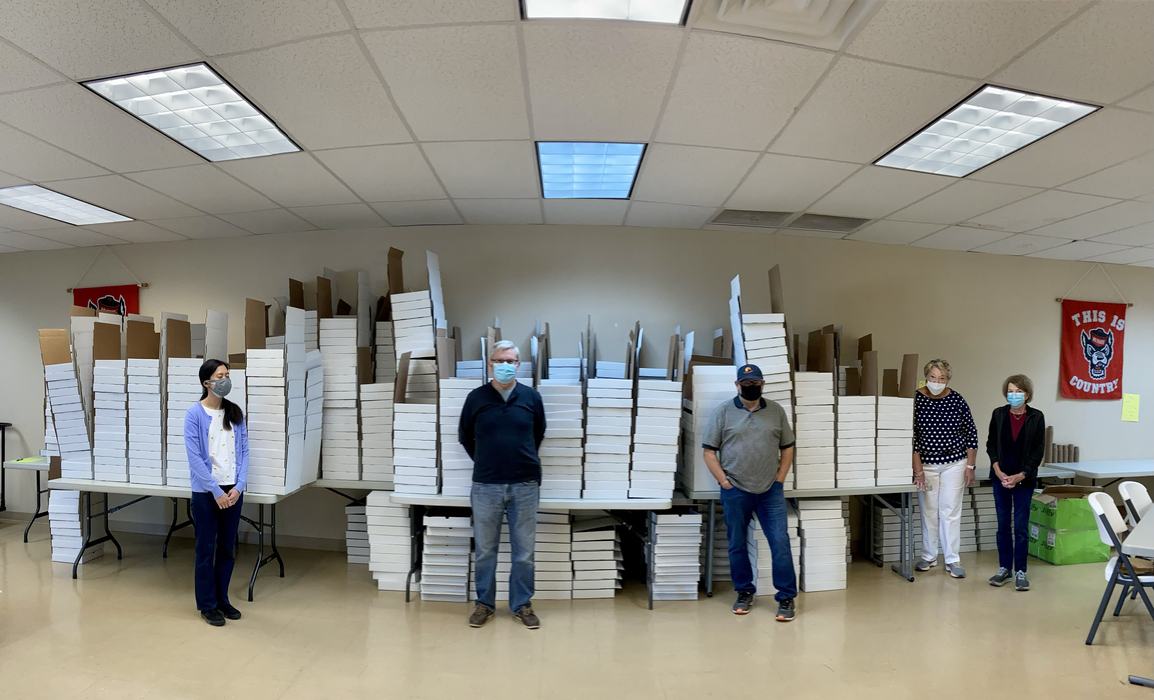 Five masked people standing spaced in front of tall stacks of white boxes in a room