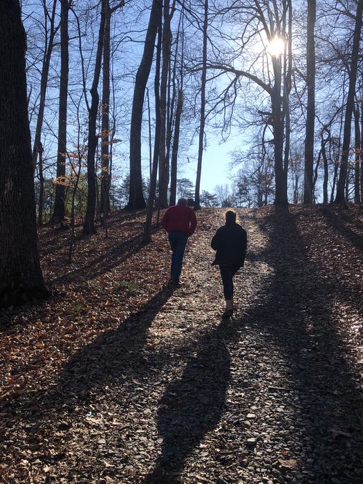Two people walking uphill on a wooded leaf-strewn path with long shadows and sun through trees