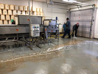 Two workers hosing a wet floor next to an industrial conveyor and stacked buckets