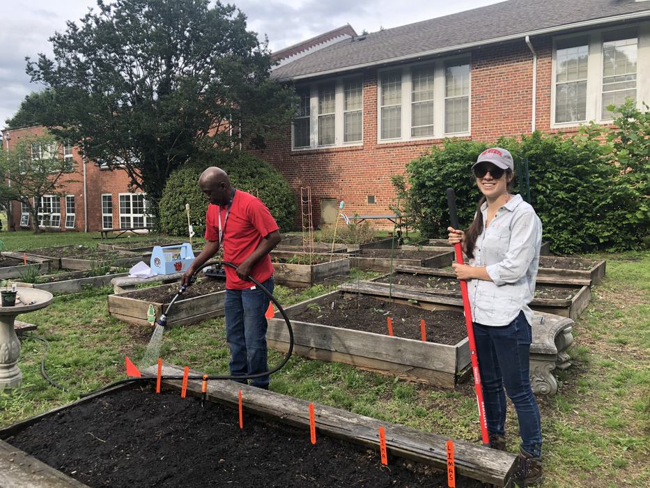 Man watering raised garden bed with hose; woman holding red shovel beside beds