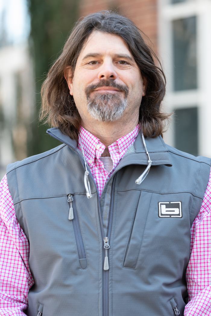 Man with shoulder-length hair and gray goatee wearing gray vest over pink checkered shirt