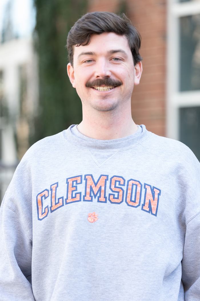 Man wearing gray sweatshirt with "CLEMSON" printed, standing outdoors