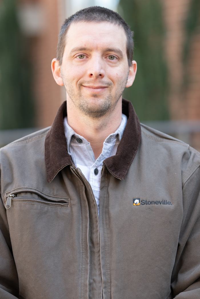 Man wearing brown jacket with "Stoneville." logo, facing camera, upper body visible