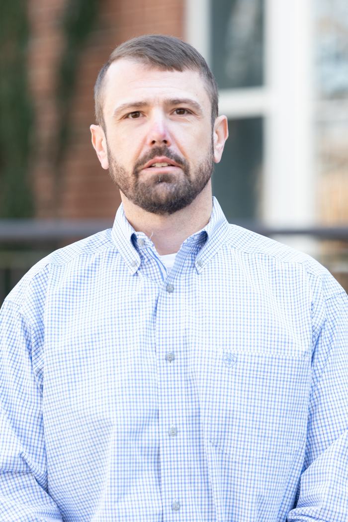 Adult man with short hair and beard wearing blue checkered button-up shirt, facing camera outdoors