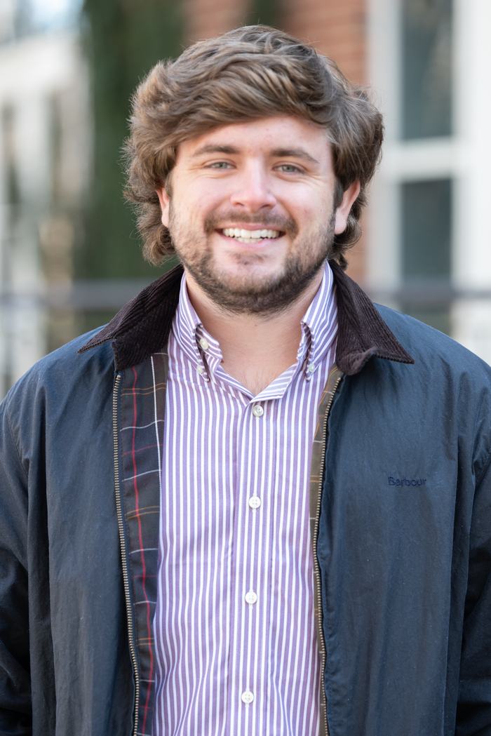 Young man wearing a purple striped button-up shirt and dark jacket, standing outdoors