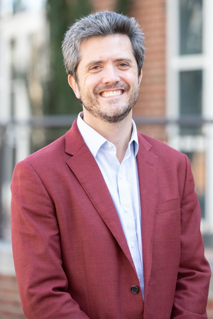 Man wearing maroon blazer and striped white shirt, smiling in outdoor portrait