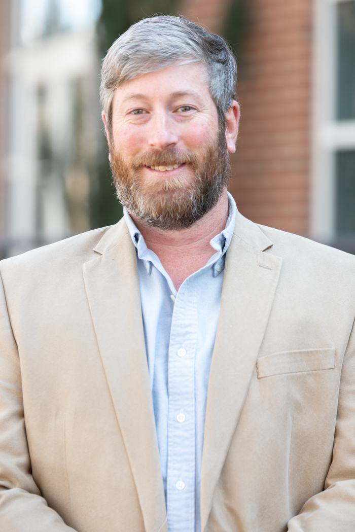Man with gray hair and beard wearing a tan blazer and light blue button-down shirt, outdoors