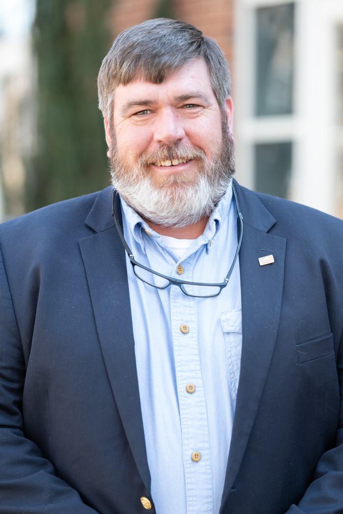 Bearded man in blazer and button-down shirt with glasses hanging around neck