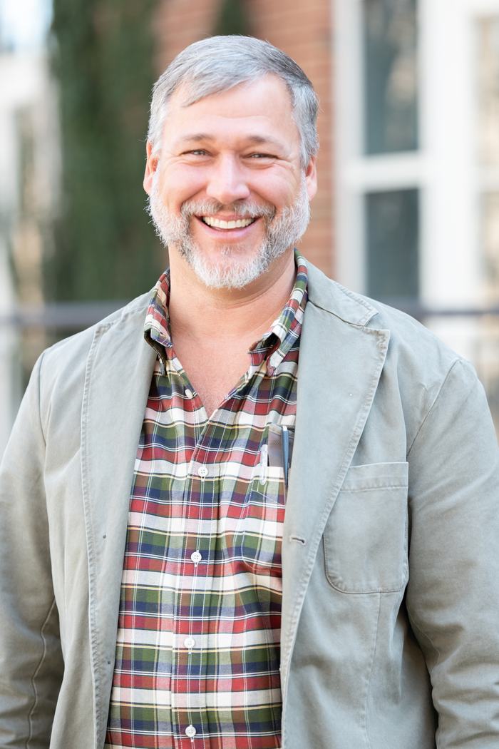 Middle-aged man smiling outdoors wearing a plaid shirt and light jacket