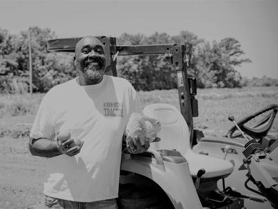 Man by tractor holding cabbage and tomatoes; shirt reads "A MAN WITH A TRACTOR"