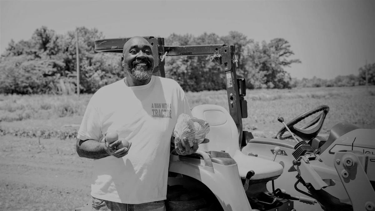 Man by tractor holding cabbage and tomatoes; shirt reads "A MAN WITH A TRACTOR"