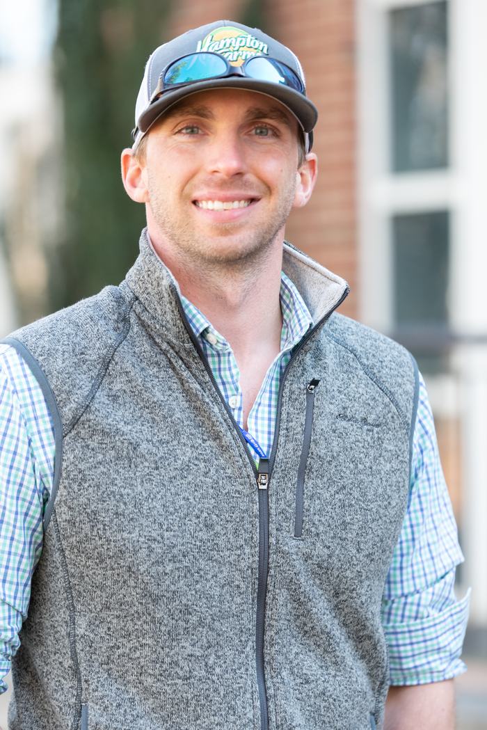 Young man wearing a cap with sunglasses on the brim, gray zip vest over plaid shirt