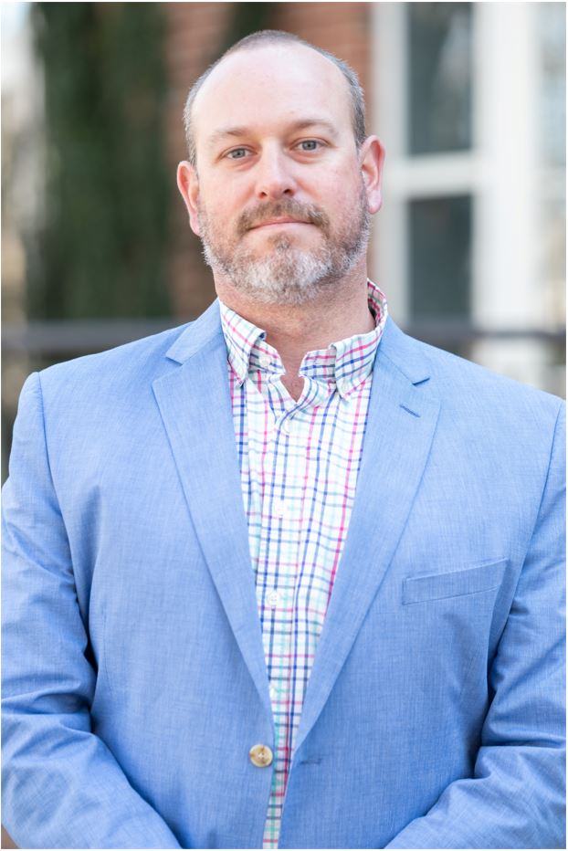Bearded man in a light blue blazer and multicolored checked shirt, facing camera.