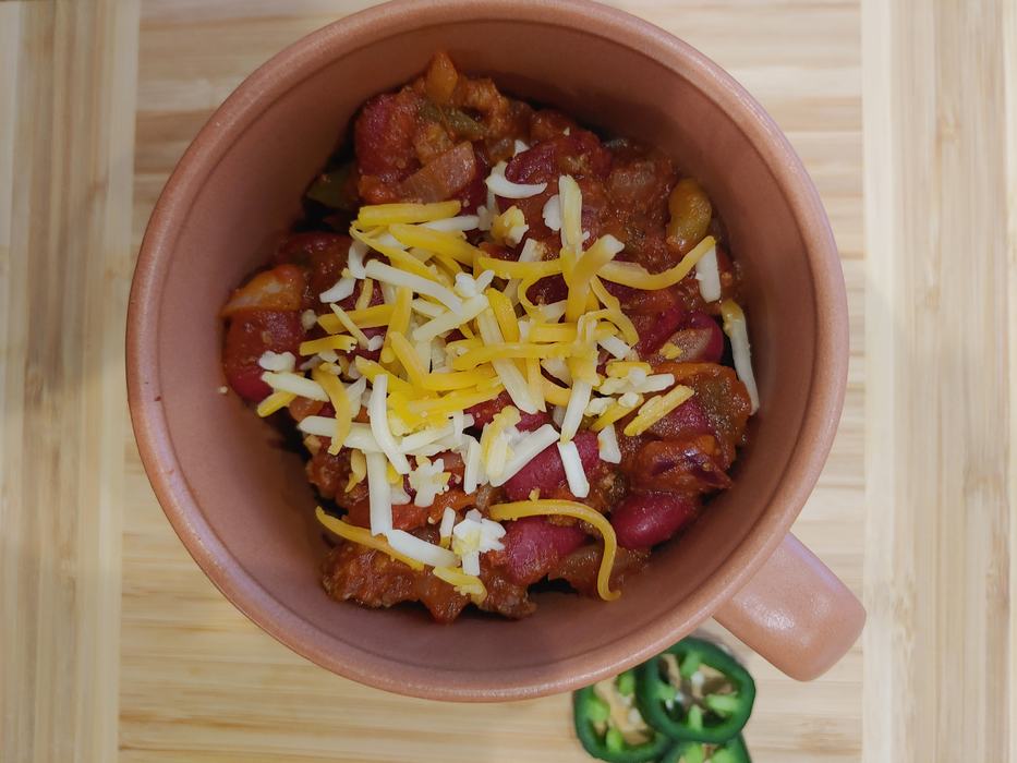 Bowl of chili with kidney beans and shredded cheddar, jalapeño slices on wooden board