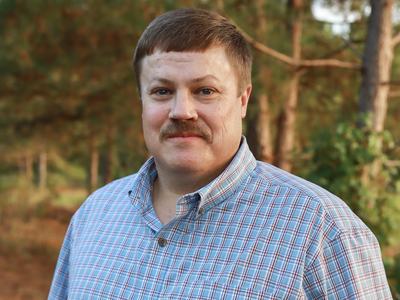 Middle-aged man with mustache in blue plaid shirt standing outdoors among pine trees.