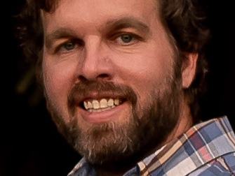 Bearded man in plaid button-down shirt, head-and-shoulders view, teeth visible.
