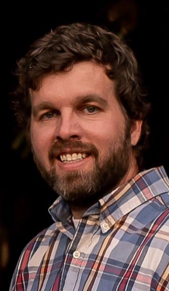 Bearded man in plaid button-down shirt, head-and-shoulders view, teeth visible.
