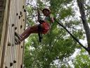 Child wearing helmet and harness rappelling down a wooden climbing wall among trees