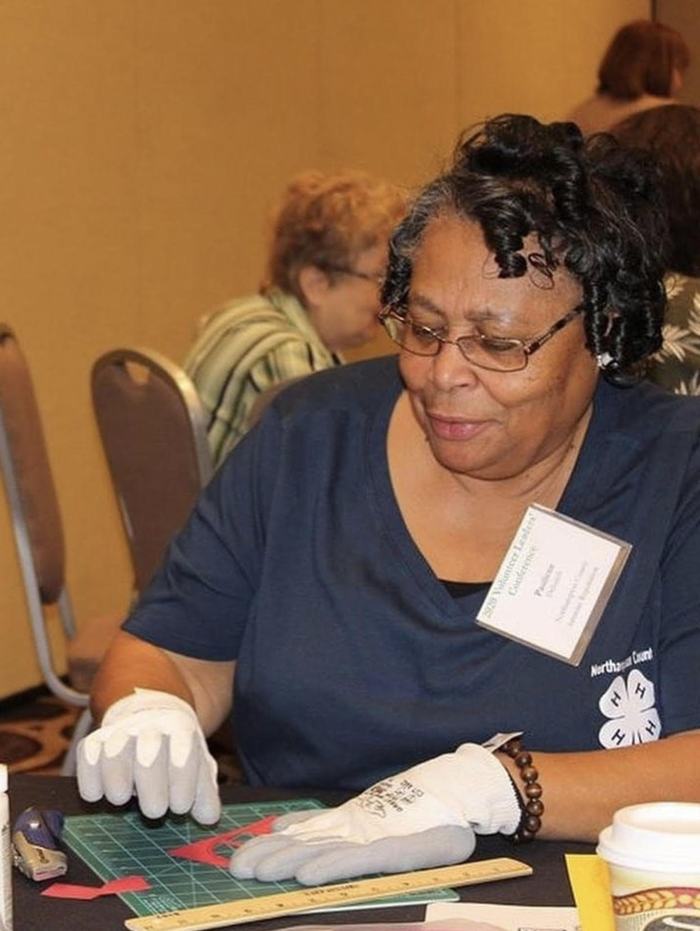 Woman wearing gloves cutting paper on a green cutting mat, ruler and 4‑H logo on shirt