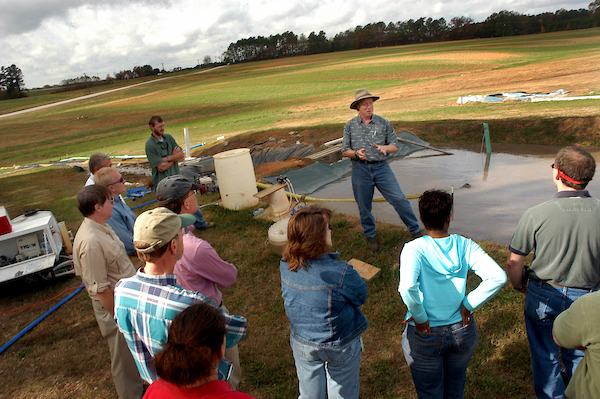 Man in hat speaking to a group beside a lined pond on a grassy field