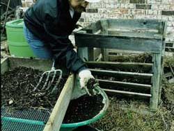Person in white cowboy hat using garden fork to load soil into green wheelbarrow by wooden sifter